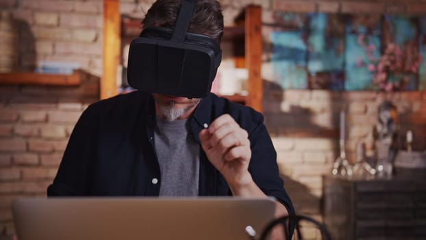 Casual man man working with virtual reality googles at home sitting at desk with laptop computer in home office.