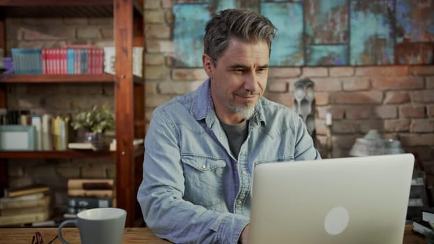 Mature man working alone from home. Sitting at desk teleworking online with laptop in home office, using internet.