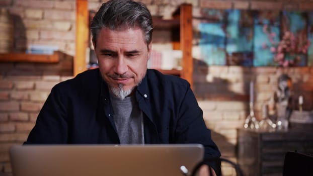 Mature adult man in 50s working on laptop in home office, working from home online, using internet, thinking.