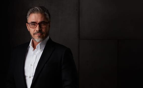 Business portrait of older man in 50s with gray hair against dark background. Businessman in glasses, copy space.