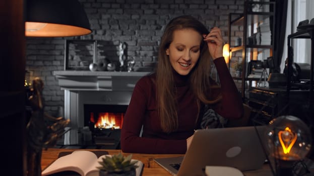 Home office - woman working from home with laptop computer. Talking on video chat, sitting at desk in dark living room in front of fireplace in Winter.
