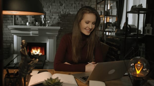 Home office - white woman working from home with laptop computer. Sitting at desk in dark living room in front of fireplace in Winter.