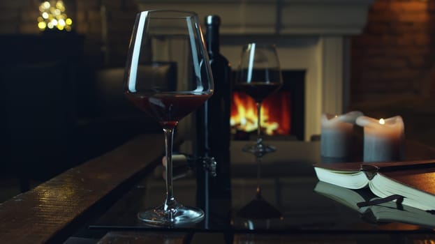 Glass of red wine, book and candle on table in dark room with fireplace. Cozy warm home in winter.