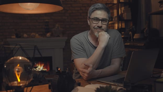 Home office - older white man working from home. Thinking, typing on laptop computer. Sitting at desk in dark living room iin front of fireplace.