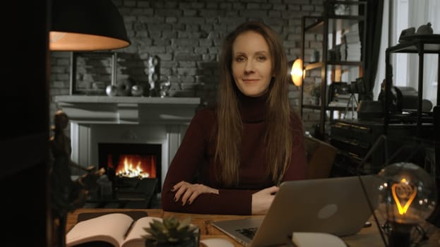 Home office - white woman working from home with laptop computer. Sitting at desk in dark living room in front of fireplace in Winter.