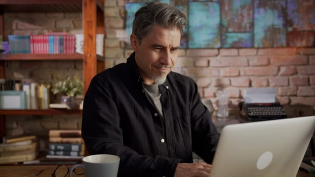 Mature man working from home, sitting at desk in home office using laptop, thinking, browsing internet, teleworking online.