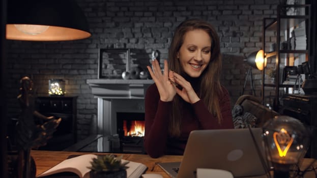 Home office - woman working from home with laptop computer. Talking on video chat, sitting at desk in dark living room in front of fireplace in Winter.