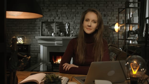 Home office - white woman working from home with laptop computer. Sitting at desk in dark living room in front of fireplace in Winter.