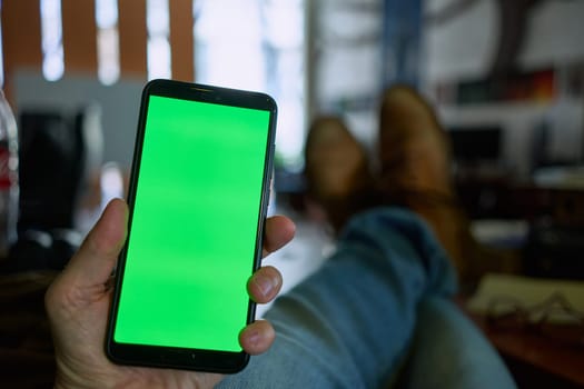 Close-up of hand holding phone with blank screen. Man sitting at desk in home office, resting with feet on table.