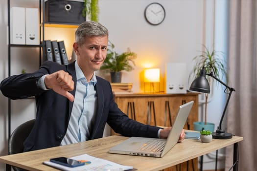 Dislike. Upset young businessman in suit working on laptop at home office showing thumb down sign gesture, disapproval, expressing discontent, dissatisfied bad work. Displeased serious male freelancer