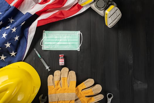 Happy Labor Day during breakout coronavirus. Several constructor work tools, labor COVID-19 vaccine, medical face mask protective and American flag with copy space on black wood background