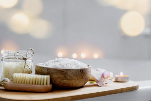 Preparation for hotel spa treatment, home bath procedure. White washbasin in bathroom, accessories on tray. Burning candles, soap, foot brush, towel, glass bottle with sea salt, orchid flower