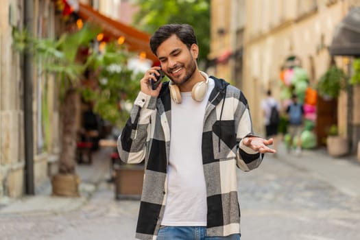 Indian young man tourist having remote conversation communicate speaking by smartphone with friend, talking on mobile phone unexpected good news gossip walking in urban city street. Town lifestyles.