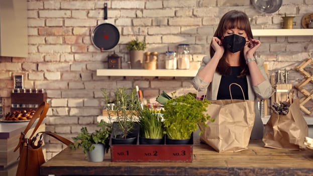 Covid-19 situation - Woman taking off face mask in the kitchen unpacking grocery bags full of vegetables arriving home from shopping.