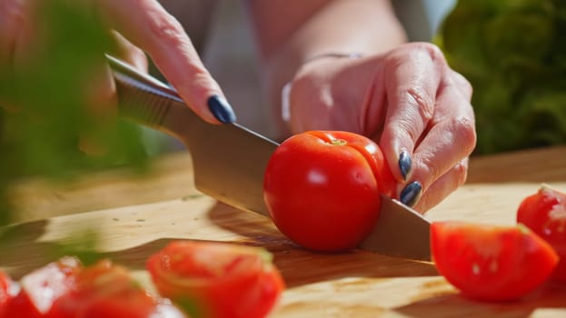 Woman slicing tomato and vegetables for cooking on kitchen table. Closeup hands. Cosy dark room.
