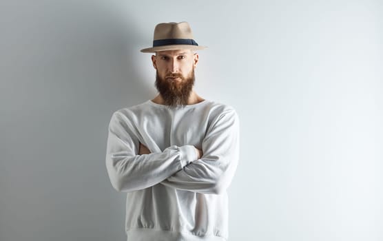 Serious bearded man in hat standing against white wall arms crossed.