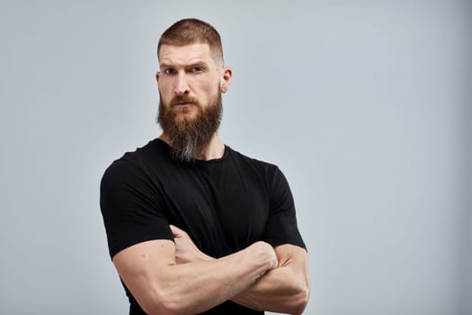 Serious muscular bearded man standing against white wall.
