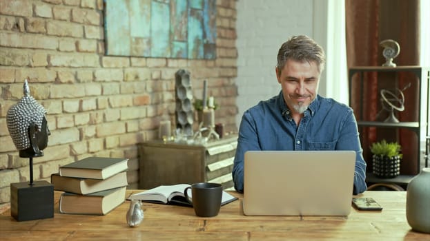 Older man tele working with laptop computer at home office in living room, thinking, looking busy.