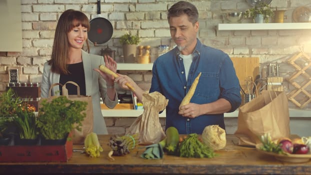 Happy couple in the kitchen unpacking grocery bags full of vegetables arriving home from shopping.