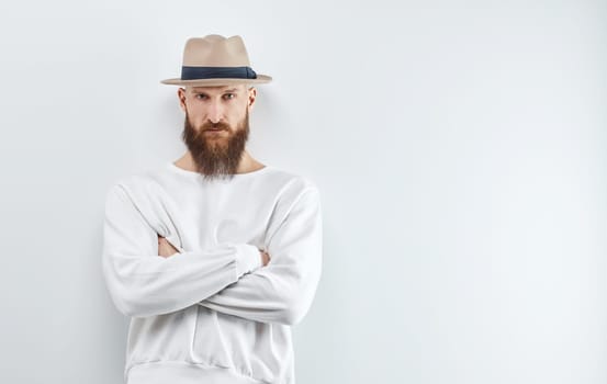 Serious bearded man ib hat standing against white wall.