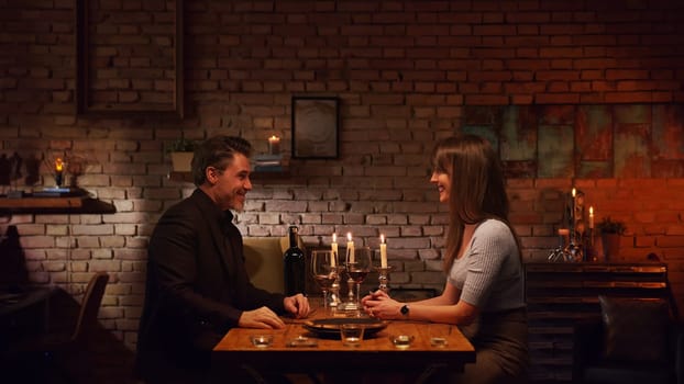 Elegant couple having romantic dinner at home, sitting at table in living room drinking red wine, talking. Stay at home concept.