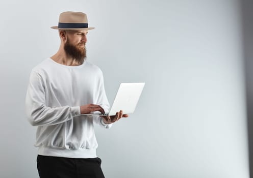 Serious bearded man in hat standing against white wall holding laptop computer.