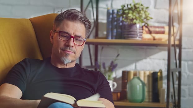 Older white man in glasses reading book at home sitting in armchair in cosy room.