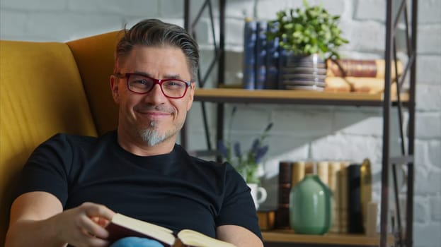 Older white man in glasses reading book at home sitting in armchair in cosy room, smiling.