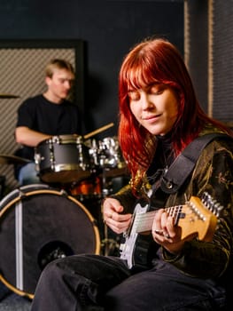 Young redhead woman playing electric guitar and a male drummer in a recording studio