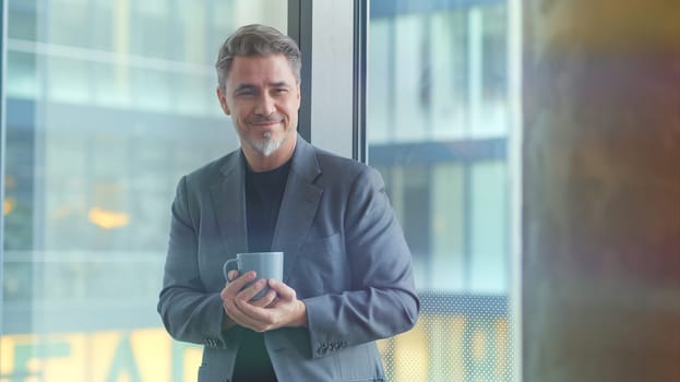 Happy older businessman taking break in office drinking coffee standing in window and daydreaming, thinking.