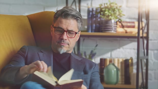 Older white man in glasses reading book at home sitting in armchair in cosy room.