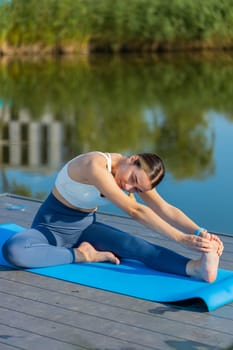 Flexible Caucasian young woman in sportswear doing stretching exercising on yoga mat near pond in park. Adult sportswoman girl bending forward represents healthy lifestyle. Athletic sporty female.