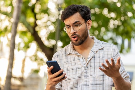 Indian young man using smartphone typing browsing, loses becoming surprised sudden lottery results bad news, fortune loss, fail. Guy standing on urban city street outdoors. Town lifestyles. Horizontal