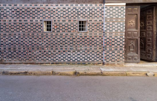 Wall with black and red bricks with white seam and two small adjacent windows with metal bars on cobblestone road, and wooden door with engraved Star of David