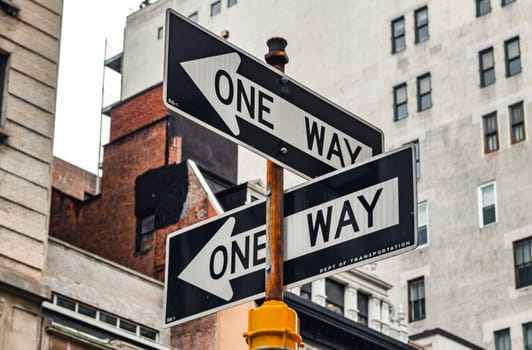 Two one way sign black colored with white arrow pointing in different directions in manhattan, new york city with building exterior in the background, view from low angle, horizontal