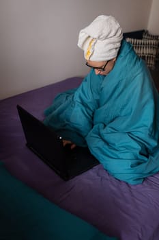 Home office, telecommuting, isolation, quarantine. A young girl working on a laptop, sitting on a bed at home in a towel after a shower