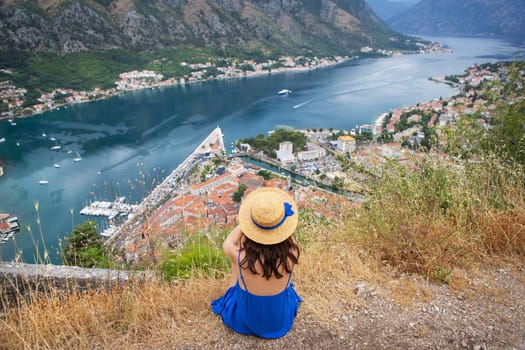 A girl in a blue dress and a straw hat sits against the backdrop of the Bay of Kotor on a beautiful summer day in Montenegro. A very beautiful fjord