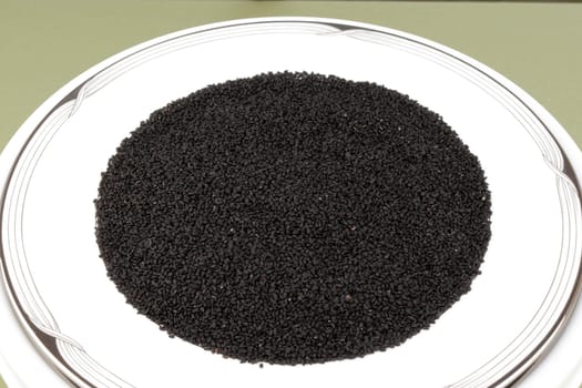 Plate full of dried raw black seed, nigella sativa food on a decorated plate close-up on a green tablecloth background. Nigella sativa, black seeds close-up on a round plate in detail of a dry food.