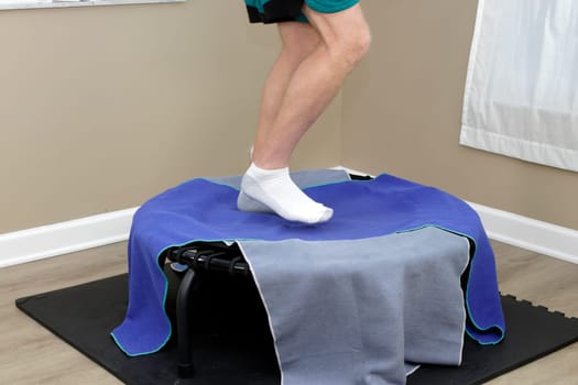 White adult male legs rebounding in a right side view on a black bungee cord rebounder with two towels covering the equipment. Walking exercising on a rebounder on an exercise mat indoors in the day.