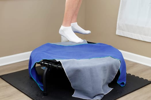Two feet in white and gray socks both bouncing up in the air on a black round bungee cord rebounder covered in two towels. Two feet in socks jumping up in the air over a round rebounder with 2 towels.