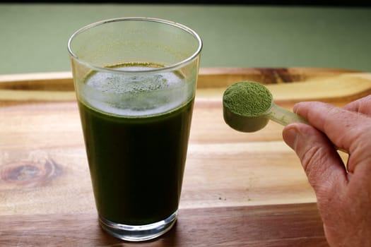 A scoop of organic celery powder being shown by fingers of a white hand near a celery powder and aloe vera juice beverage. Aloe vera juice with celery powder drink near a hand showing celery powder.