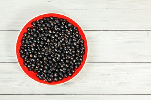 Tabletop view, red ceramic bowl full of freshly harvested blackcurrants, on white boards desk.