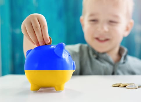 Child sitting at the table with money coins and biggybank in colour of Ukranian flag. Concept of the assistant, helping and donation for Ukraine.