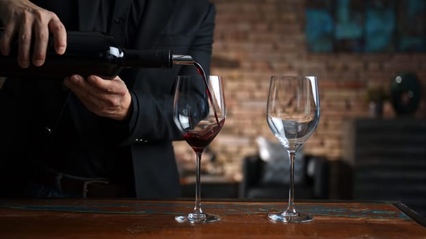 Elegant man pouring red wine from wine bottle to wine glasses at home in a cosy dark room.