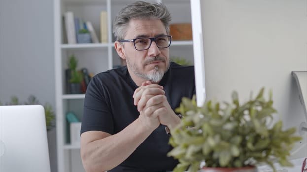 Older white man in glasses sitting at desk using desktop computer in home office. 50s businessman staying home in casual working online thinking on business solution.