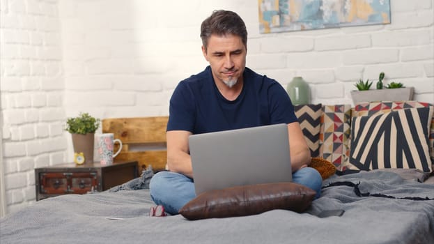 Casual man sitting on bed working online from home with laptop computer. Stay at home and work in home office.