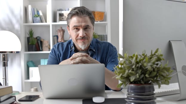 Portrait of happy older man sitting at desk working with laptop in home office. Confident 50s businessman in casual with gray hair smiling.