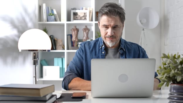 Older white man sitting at desk using laptop in home office. 50s businessman staying home in casual working online with computer thinking on business solution.