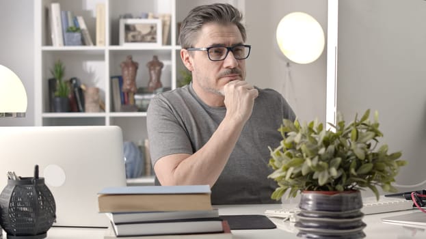 Older white man in glasses sitting at desk using laptop in home office. 50s businessman staying home in casual working online with computer thinking on business solution.