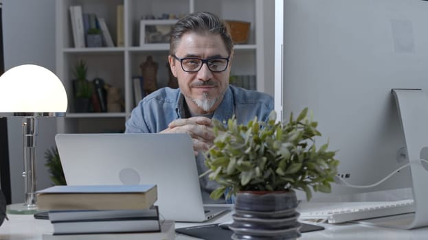 Portrait of happy older man in glasses sitting at desk working with laptop in home office. Confident 50s businessman in casual with gray hair smiling.